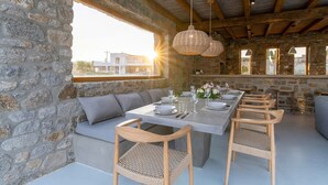 A stone-walled dining area with a long table set for six, cushioned seating, wicker chairs, and hanging wicker pendant lights. - Villa Demetra with pool - My Rental Homes (Mykonos)