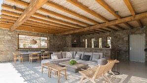 A spacious living area with a stone wall, a wooden ceiling, and a large window overlooking a scenic landscape. - Villa Demetra with pool - My Rental Homes (Mykonos)