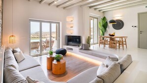 A modern living room with a sofa, coffee table, TV, and a dining area with a table and chairs. - Villa Demetra with pool - My Rental Homes (Mykonos)
