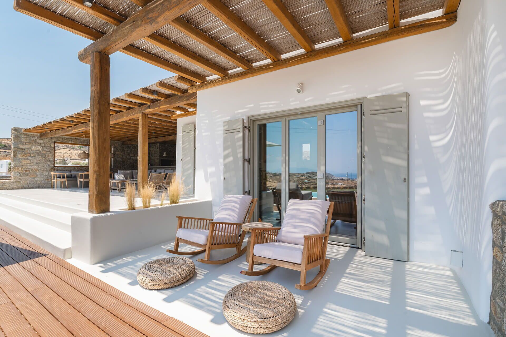 A modern outdoor seating area with wooden chairs and woven ottomans, a stone wall, and a view of the landscape.