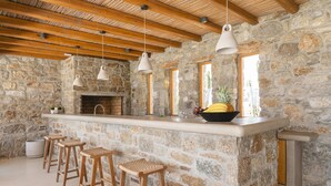 A stone-walled kitchen with a wooden ceiling, a built-in bar with stools, and a fruit bowl on the counter. - Villa Demetra with pool - My Rental Homes (Mykonos)
