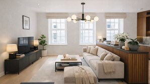 A modern living room with a sofa, coffee table, and TV. There are windows, a plant, and a kitchen area visible. - Sophisticated 3-Bed Residence for 6 | Urban Calm (London)