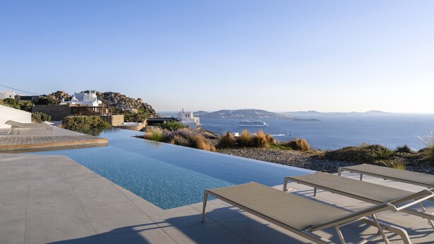 Ein Infinity-Pool mit Liegestühlen, Meerblick und einer felsigen Hügelkuppe mit Gebäuden.