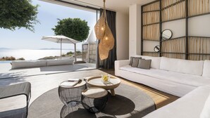 Ein modernes Wohnzimmer mit einem weißen Sofa, einem runden Couchtisch und Korbstühlen. Ein großes Fenster bietet Blick aufs Meer.