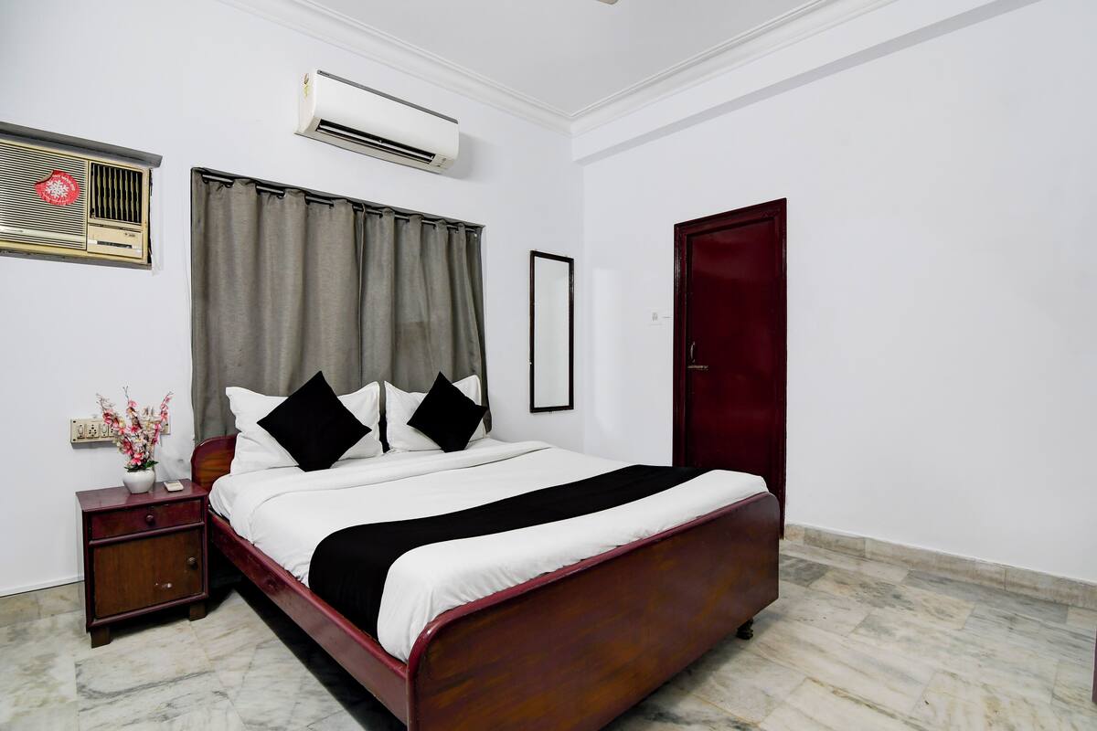A neatly arranged bedroom with a bed, a nightstand, an air conditioning unit, a mirror, and a door.