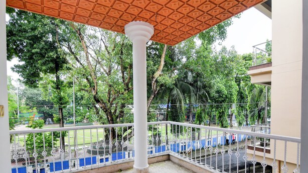 A balcony with a white railing, a central column, and a view of a green park with trees and a playground. - Smart stay by- Brill rooms (Barasat)