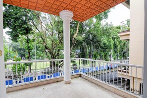 Ein Balkon mit weißer Reling, einer zentralen Säule und Blick auf einen grünen Park mit Bäumen und einer Spielplatz.