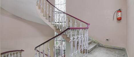 A staircase with ornate white railings and a red handrail, leading to a landing with a fire extinguisher mounted on the wall.