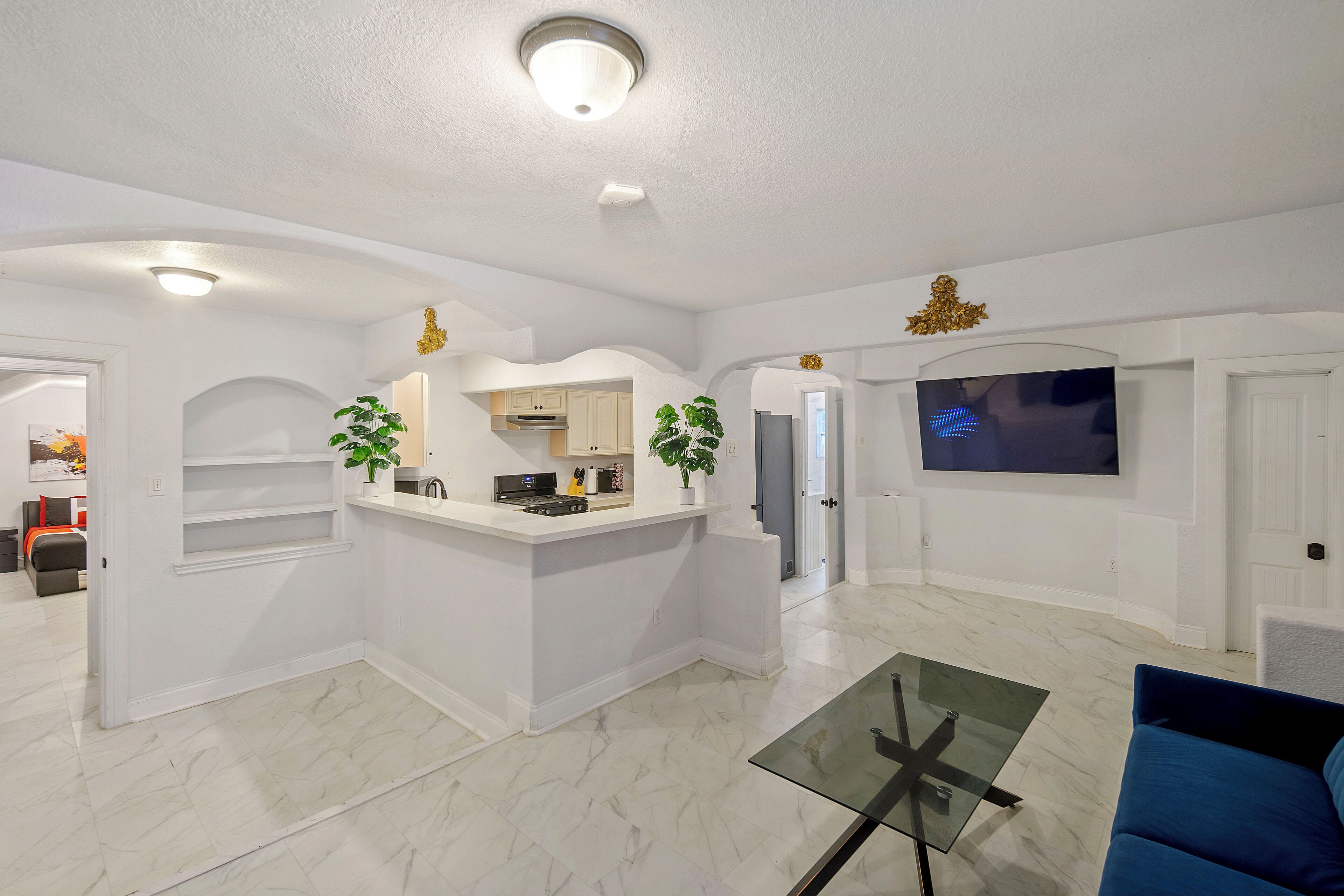 A modern living room with a glass coffee table, a TV mounted on the wall, and a kitchen area with white cabinetry.