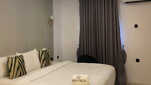 A modern hotel room with a neatly made bed, a ceiling light, a wall-mounted air conditioner, and a window with curtains. - GREYFIELD APARTMENT 2 (Benin City)