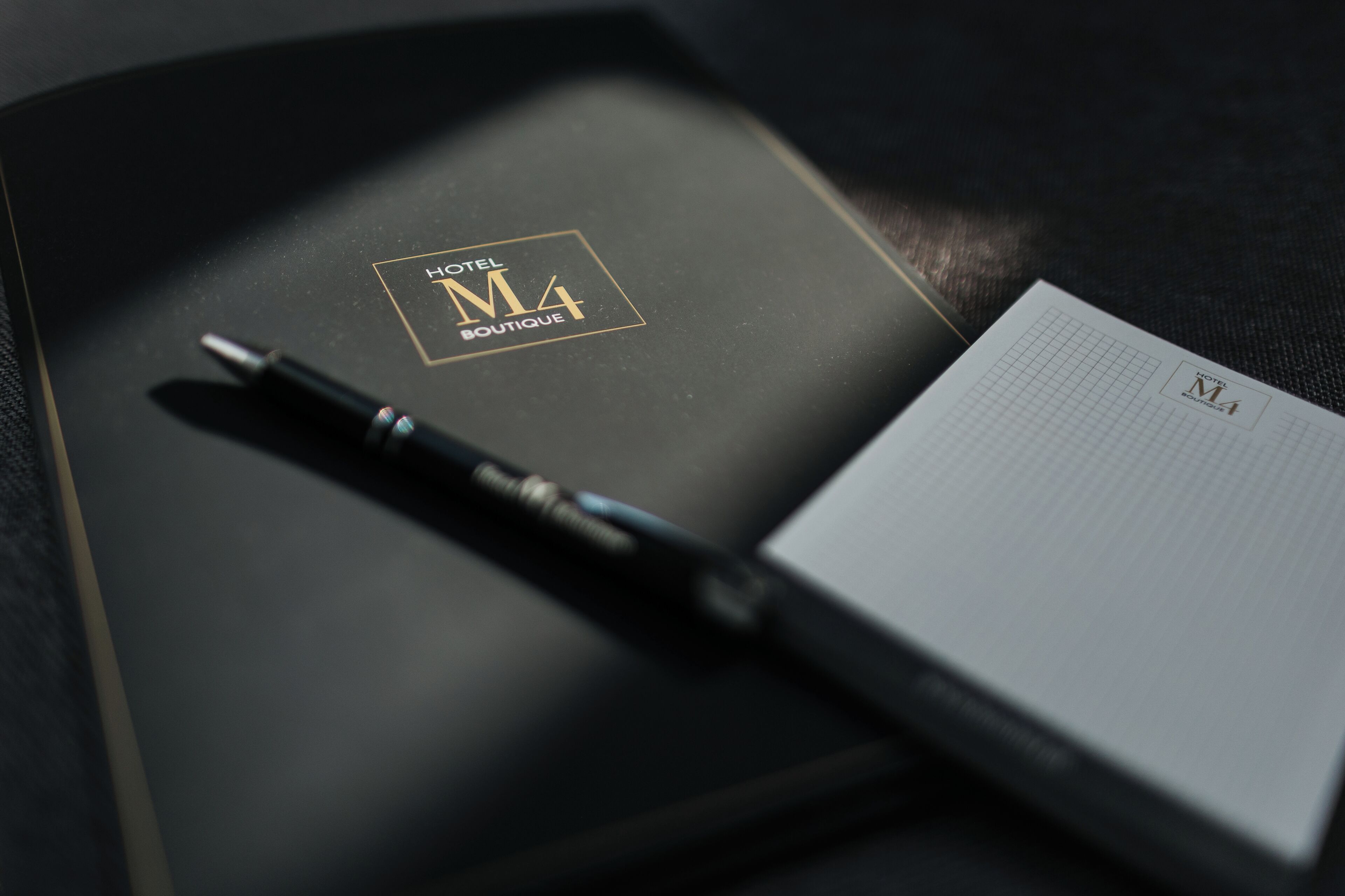 A black notebook with a pen, a grid notebook, and a black folder with 'HOTEL M4 BOUTIQUE' text.