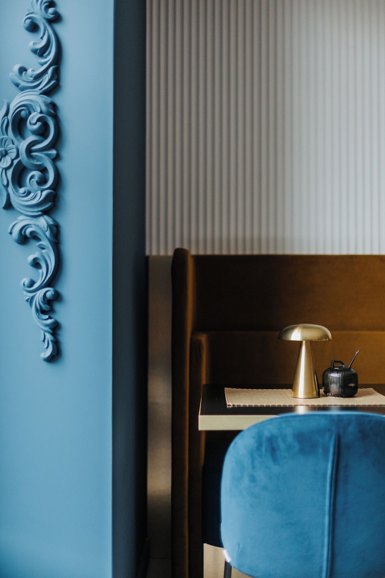 A room with a blue wall, a decorative wall piece, a bed with a headboard, a bedside table with a lamp, and a small black object on the table.