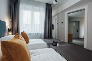 A hotel room with two beds, a bedside table, a lamp, a window with curtains, and a door leading to another room.