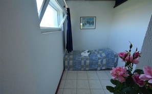 A small, narrow room with a single bed, a window with a curtain, a framed picture on the wall, and a potted plant with pink flowers. - Residence With Pool in Lignano Riviera (Lignano Sabbiadoro)