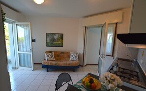 A compact living space with a sofa, a dining table with fruit, a kitchen area, and a painting on the wall. - Residence With Pool in Lignano Riviera (Lignano Sabbiadoro)