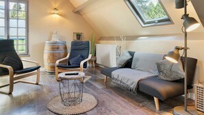 Un acogedor espacio tipo loft con techo inclinado, un ventanal grande, un sofá, una silla, una mesita redonda y una lámpara de pie.