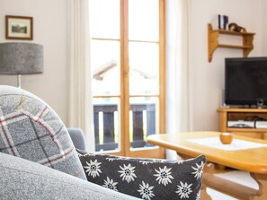 Ein Wohnzimmer mit einer Couch, einem Tisch, einem Fernseher und einem Fenster mit Blick ins Freie.