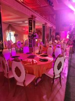 Salão de banquetes com mesas redondas, cadeiras brancas com detalhes dourados e um arranjo floral central.