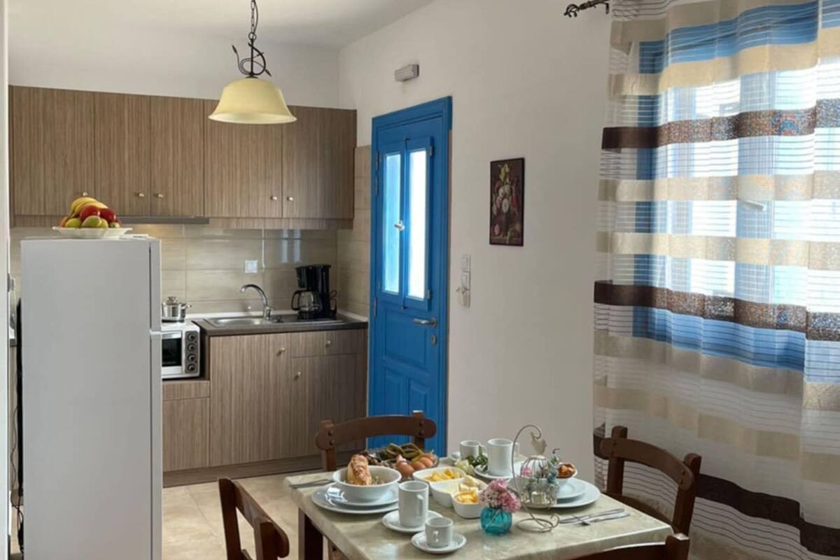Cocina compacta con refrigerador, microondas y gabinetes de madera. Una puerta azul da acceso al área de comedor, con una mesa lista para dos comensales.