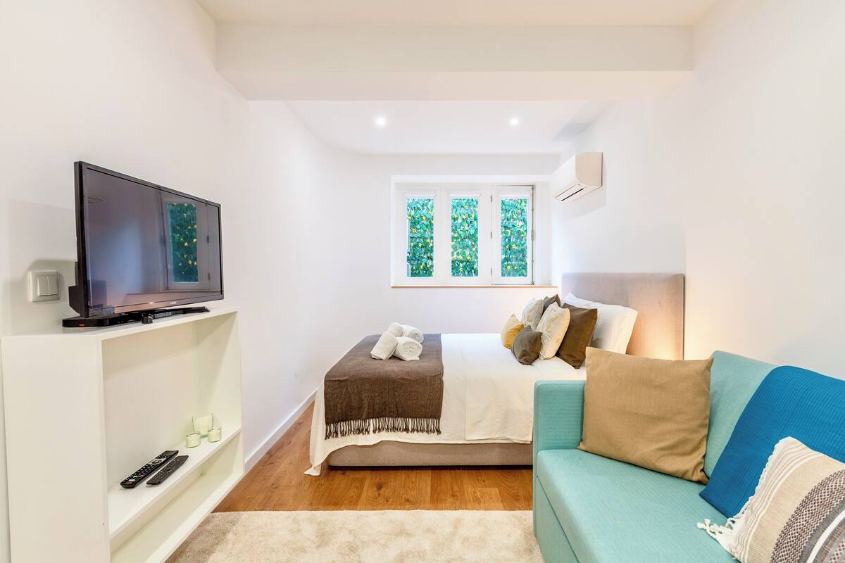 A modern bedroom with a bed, a TV on a stand, a window with a view of greenery, and a blue sofa.