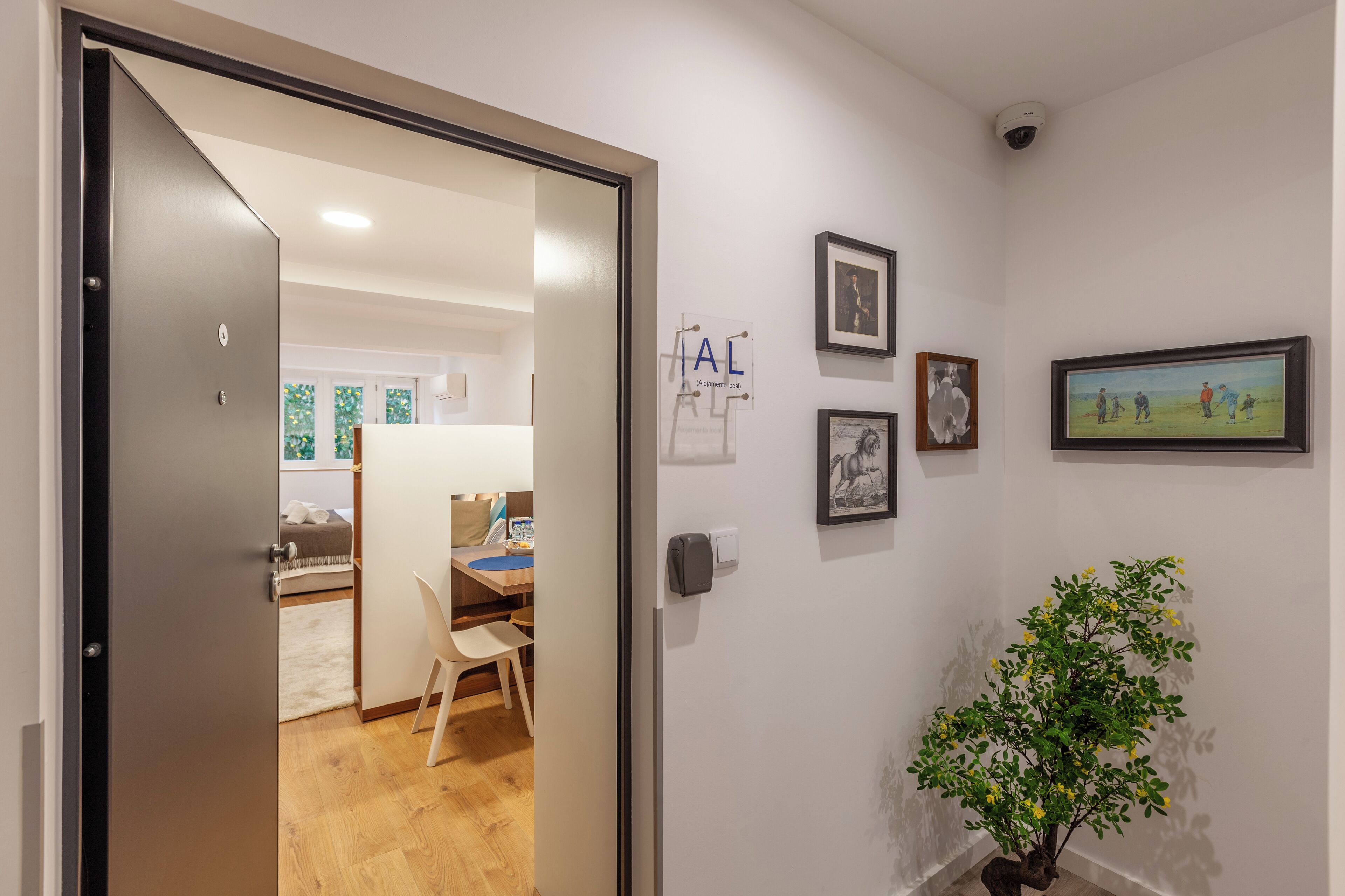 Un pasillo con una puerta que da a una habitación con una cama y un escritorio. En la pared hay cuadros enmarcados y una pequeña planta en una maceta.