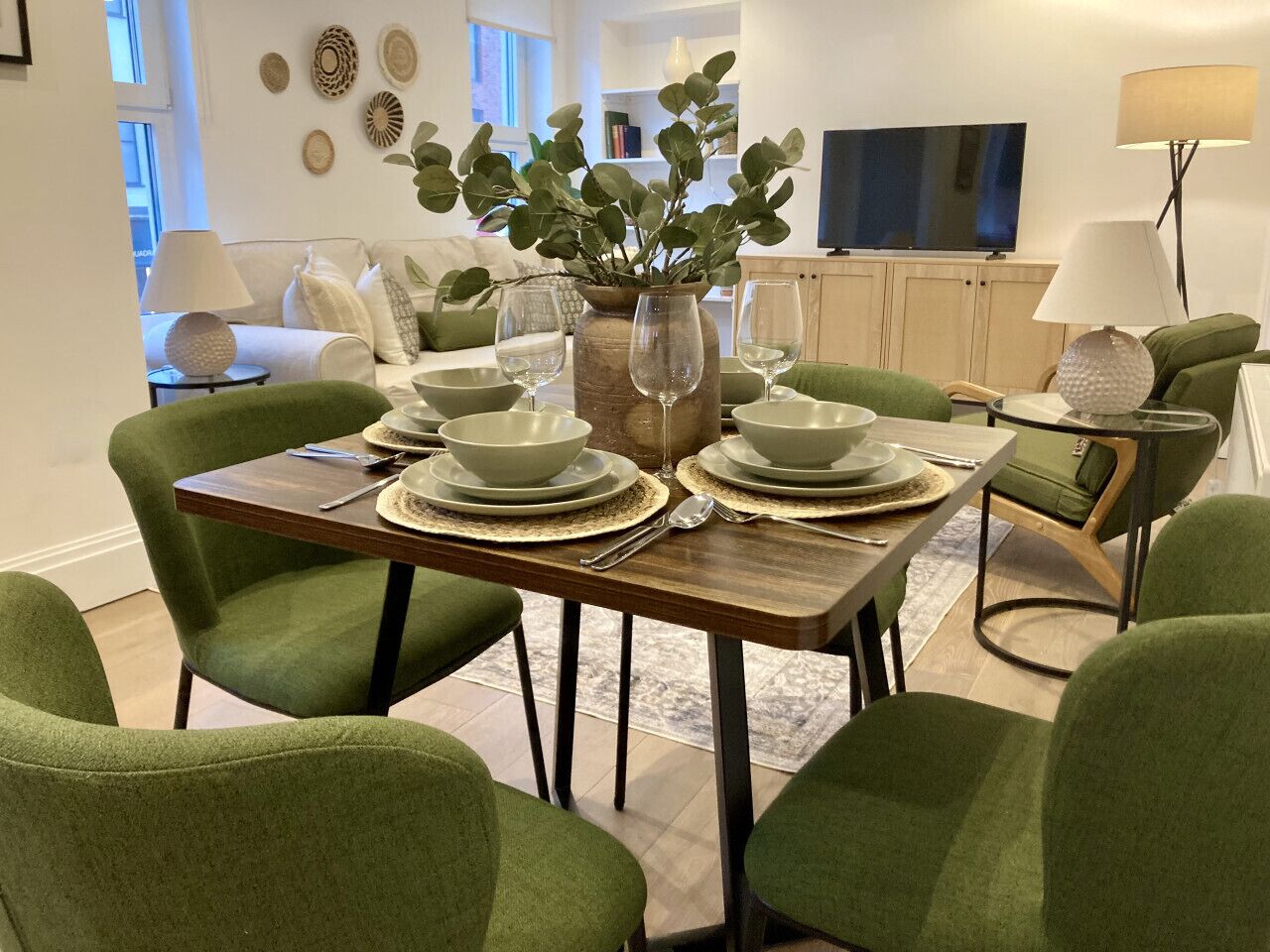 Ein Essbereich mit einem Holztisch, gedeckt für vier Personen, umgeben von grünen, gepolsterten Stühlen. Auf dem Tisch steht eine große Vase mit Grünpflanzen.