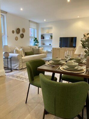 A modern living room with a dining area, featuring a sofa set, a coffee table, and a TV in the background. - Richmond High Street Flat – Station Steps Away (Greater London)