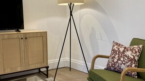 Una sala de estar moderna con un televisor de pantalla plana sobre un mueble de madera, una lámpara de pie, un sillón verde con un cojín estampado y una mesa de centro de vidrio.