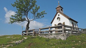 A small white church with a bell tower, surrounded by a stone wall and a wooden fence, set on a grassy hill. - Cozy vacation home in charming nature and mountain scenery! Dogs welcome! (Abtenau)