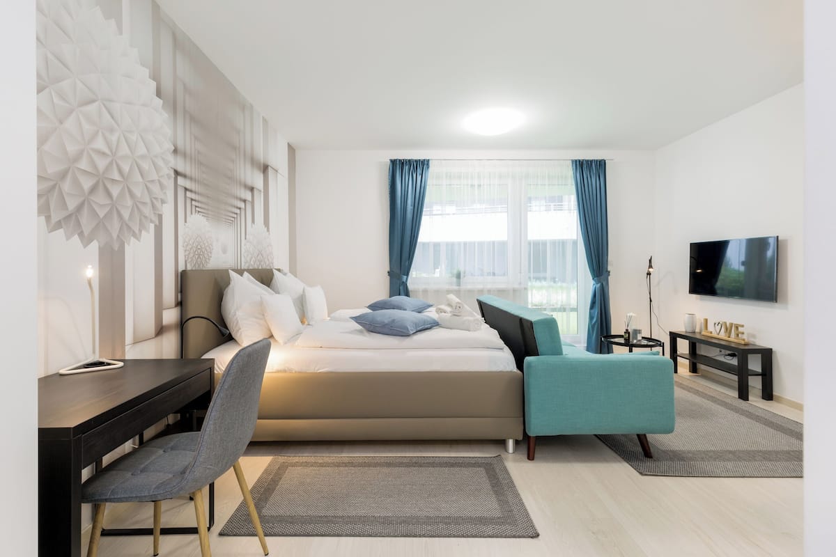 Un dormitorio moderno con cama, escritorio, silla, televisor y ventana con cortinas.