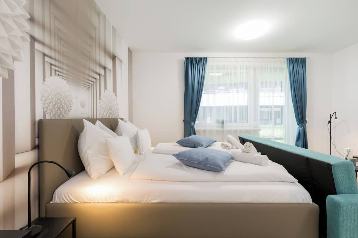 Habitación de hotel moderna con una cama grande, una mesita de noche, una lámpara y vistas al exterior a través de una ventana con cortinas azules.