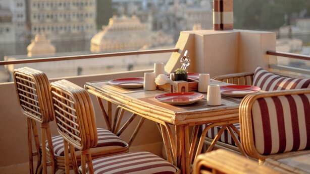 A dining area with wicker chairs and a table set for two, overlooking a cityscape with buildings and a mountain in the distance. - Bun And Bunks Udaipur (Udaipur)