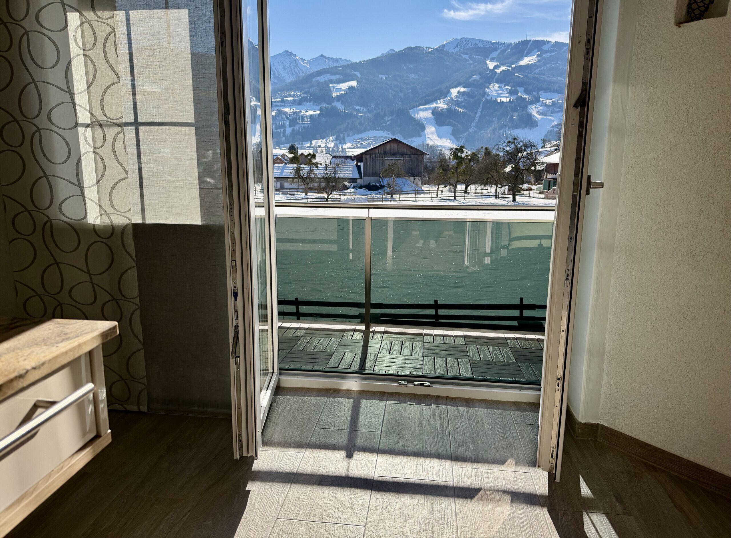 A room with a large glass door leading to a balcony with a view of a snowy mountain and a body of water.