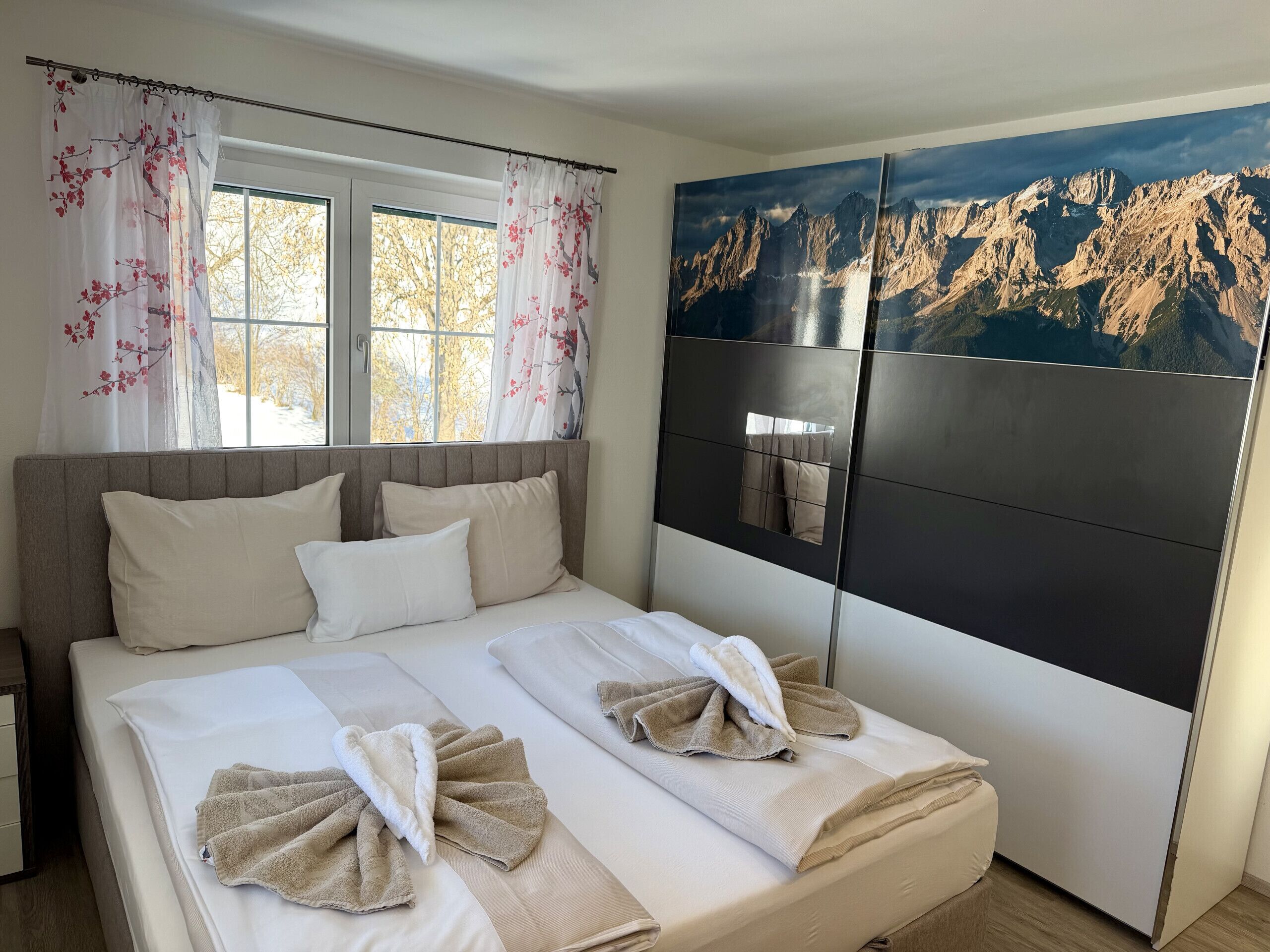 A hotel room with a bed, two windows with floral curtains, a large painting of mountains, and a bedside table.
