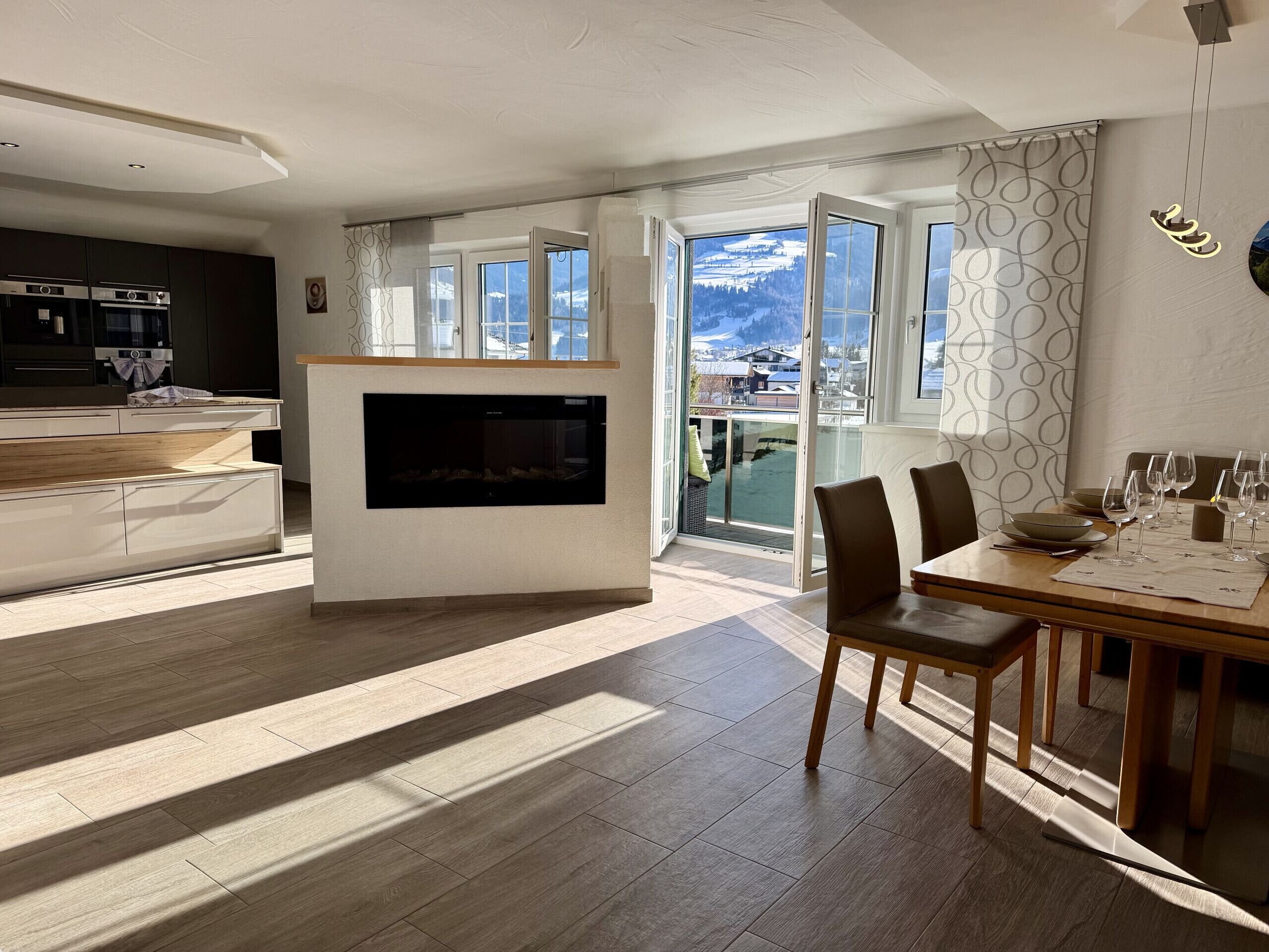 A modern kitchen with a built-in oven and microwave, a dining area with a wooden table and chairs, and a large window offering a view of a snowy landscape.