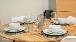 Una mesa de comedor redonda de madera dispuesta con platos, tazones y copas de vino blancos, rodeada de sillas blancas.