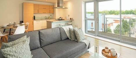 En moderne stue med en grå sofa, et glasbord og udsigt over byen gennem store vinduer.