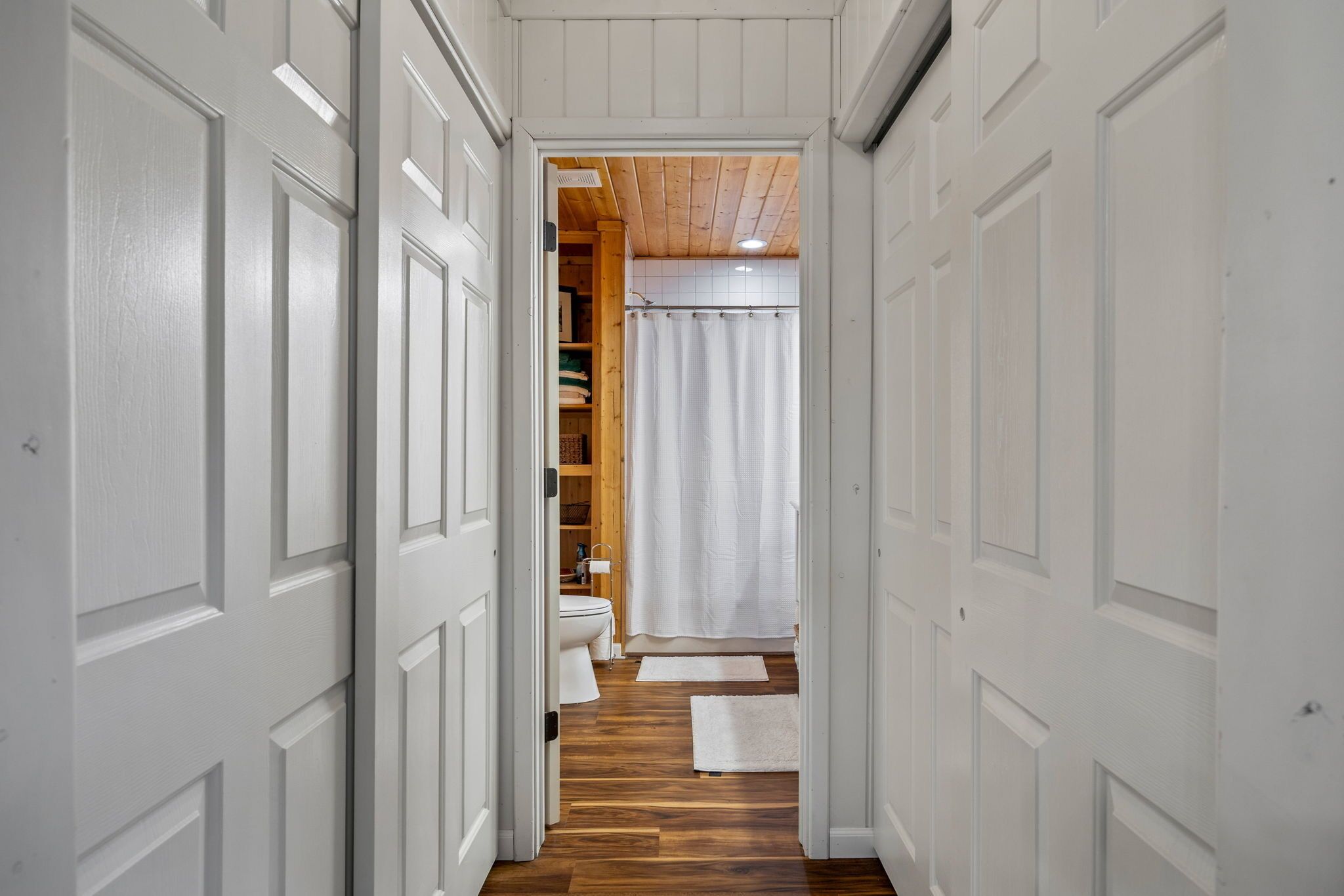 Un pasillo estrecho con suelo de madera y puertas blancas que dan a un baño con cortina de ducha.