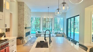 A modern kitchen with a large island, stone wall, and large windows offering a view of the forest. - FIFA World Cup. Brand new 6 BD, 8 BA, Whiskey Room, Games room+ more (West Vancouver)