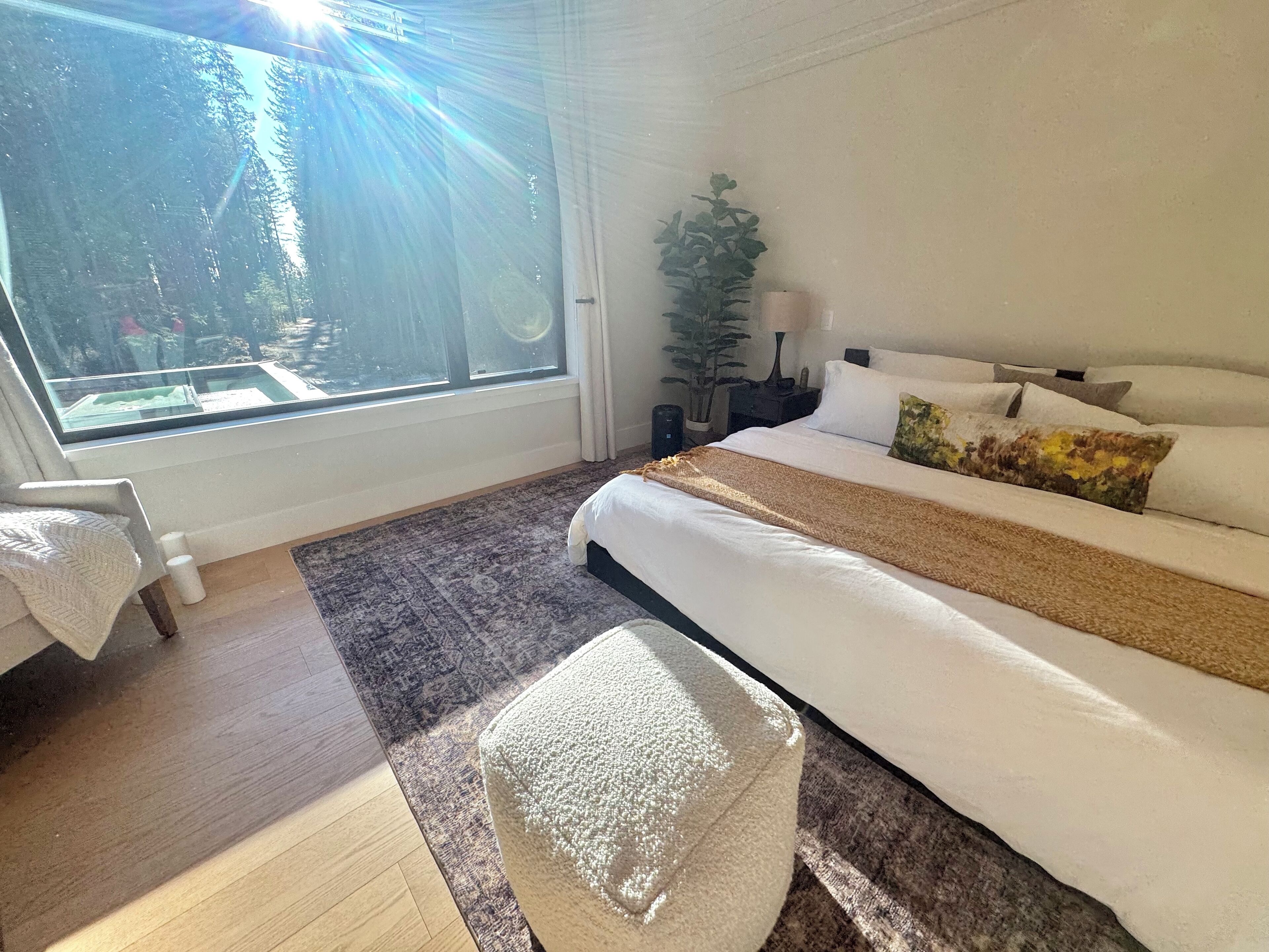 Ein Schlafzimmer mit großem Fenster, einem Bett mit weißer Bettwäsche, einem kleinen Nachttisch und einem Topfpflanzen.