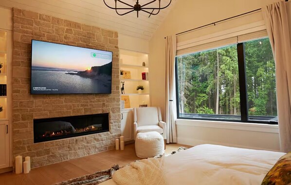 Un dormitorio moderno con chimenea de piedra, un televisor grande montado en la pared, un sillón cómodo y un ventanal con vistas al bosque.