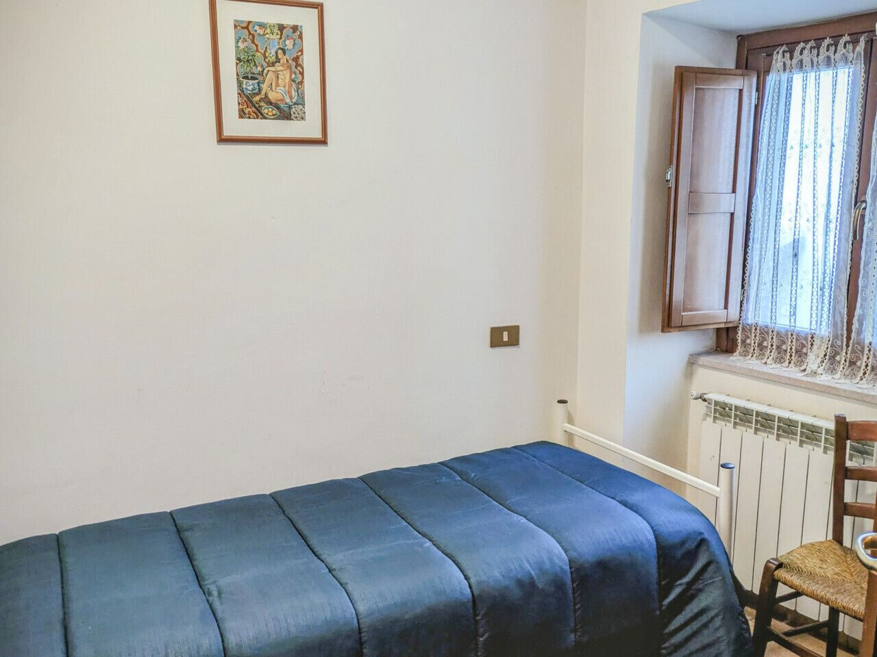 Una habitación pequeña con cama individual, colcha azul, una silla de madera, ventana con cortinas de encaje y un cuadro en la pared.