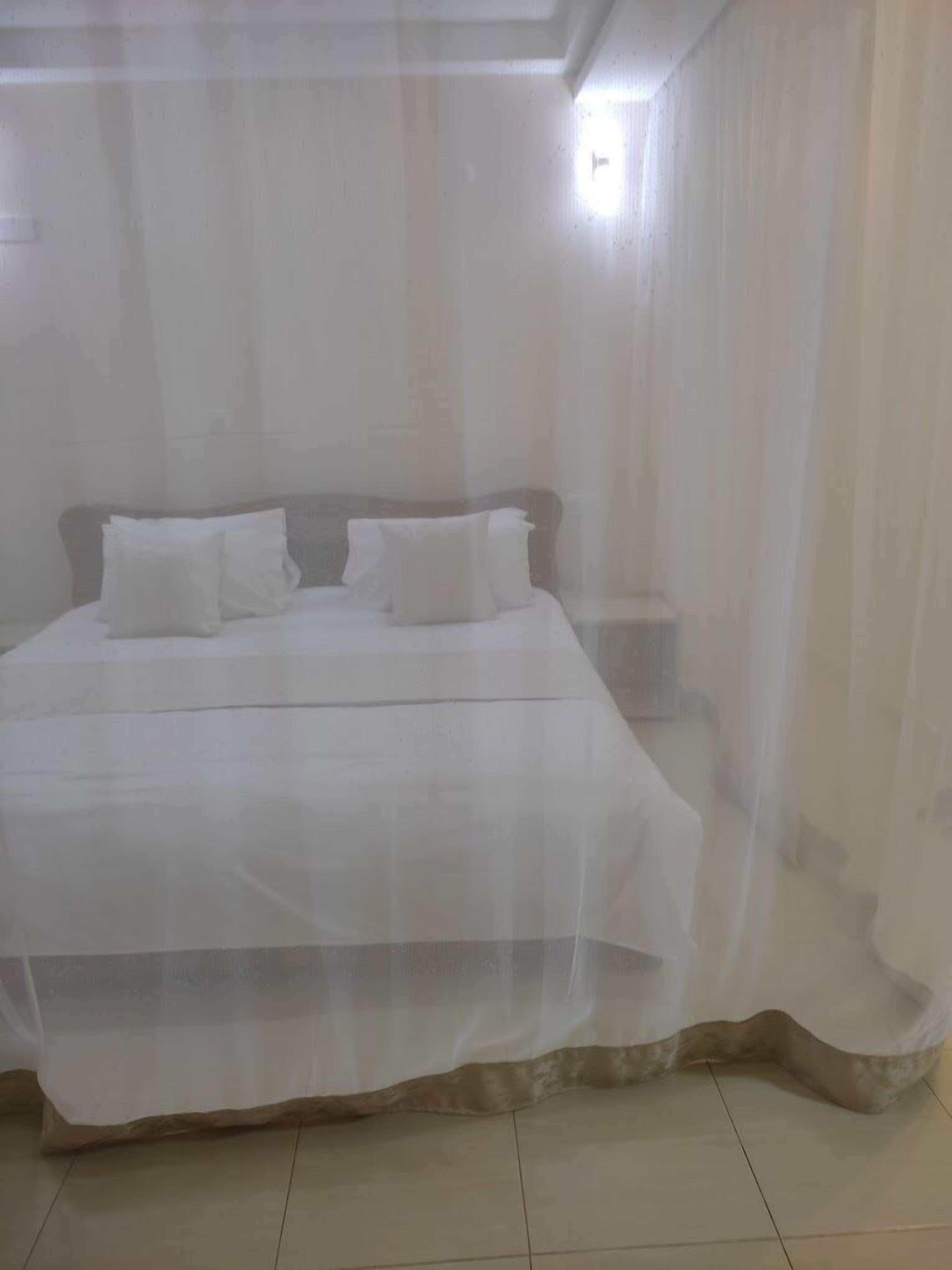 A bed with white bedding and pillows, a wooden headboard, and a sheer white curtain.