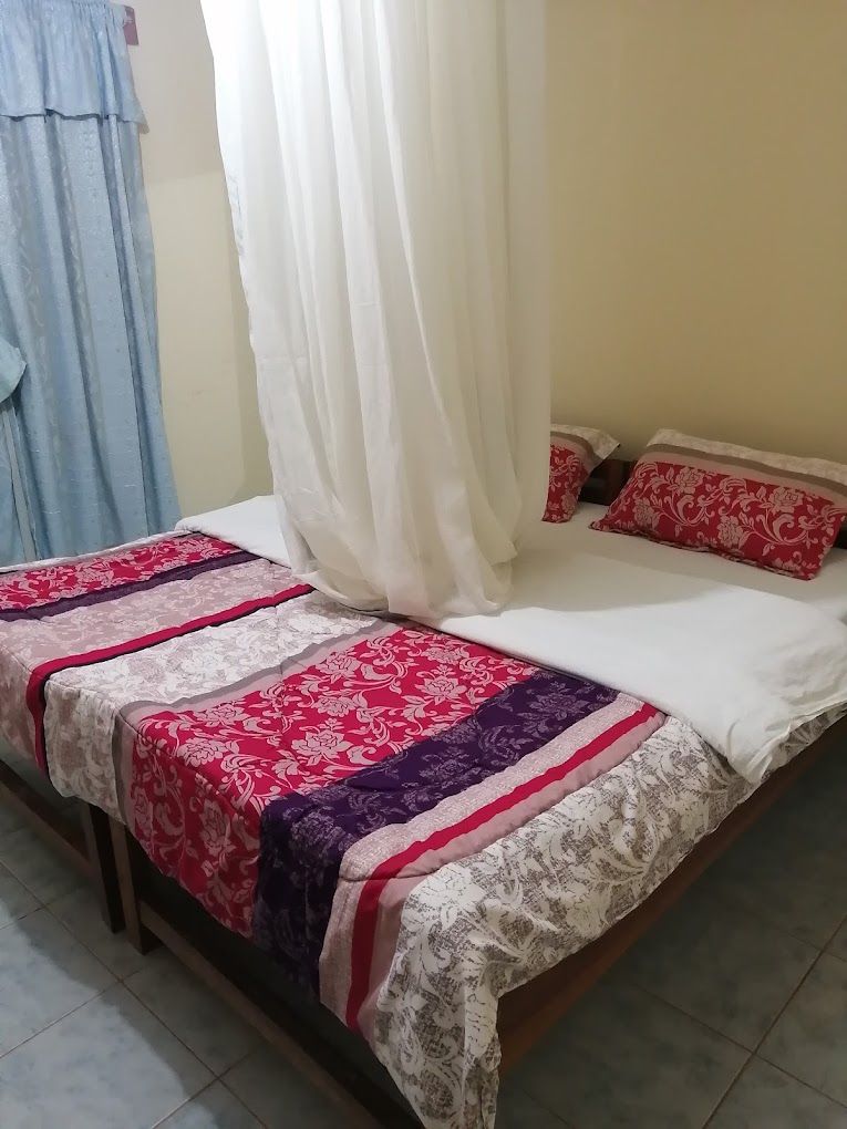 Un lit avec un drap blanc et deux oreillers, une couverture à motifs rouges et blancs, et un rideau bleu.