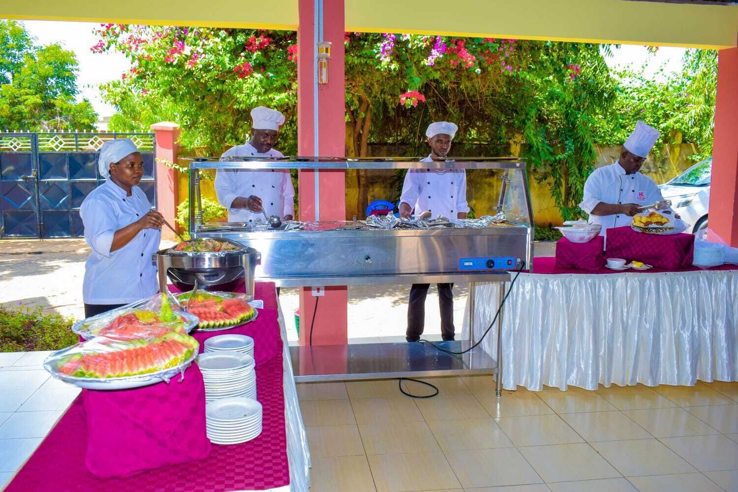 A buffet setup with chefs preparing food, a stainless steel grill, and a variety of dishes on a table covered with a pink tablecloth.