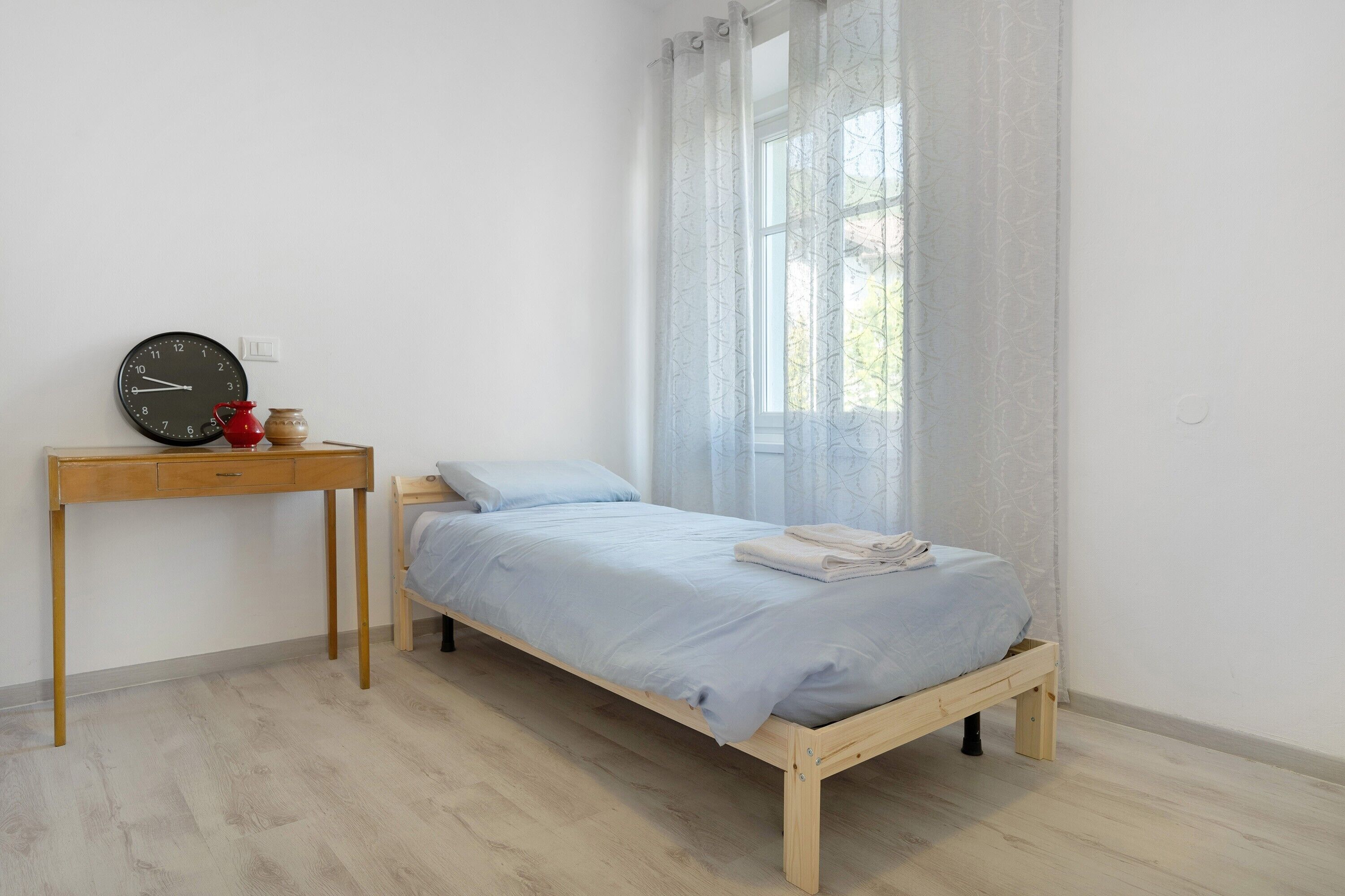 Ein Schlafzimmer mit einem Bett, einem Holztisch, einer Uhr und einem Fenster mit Vorhängen.