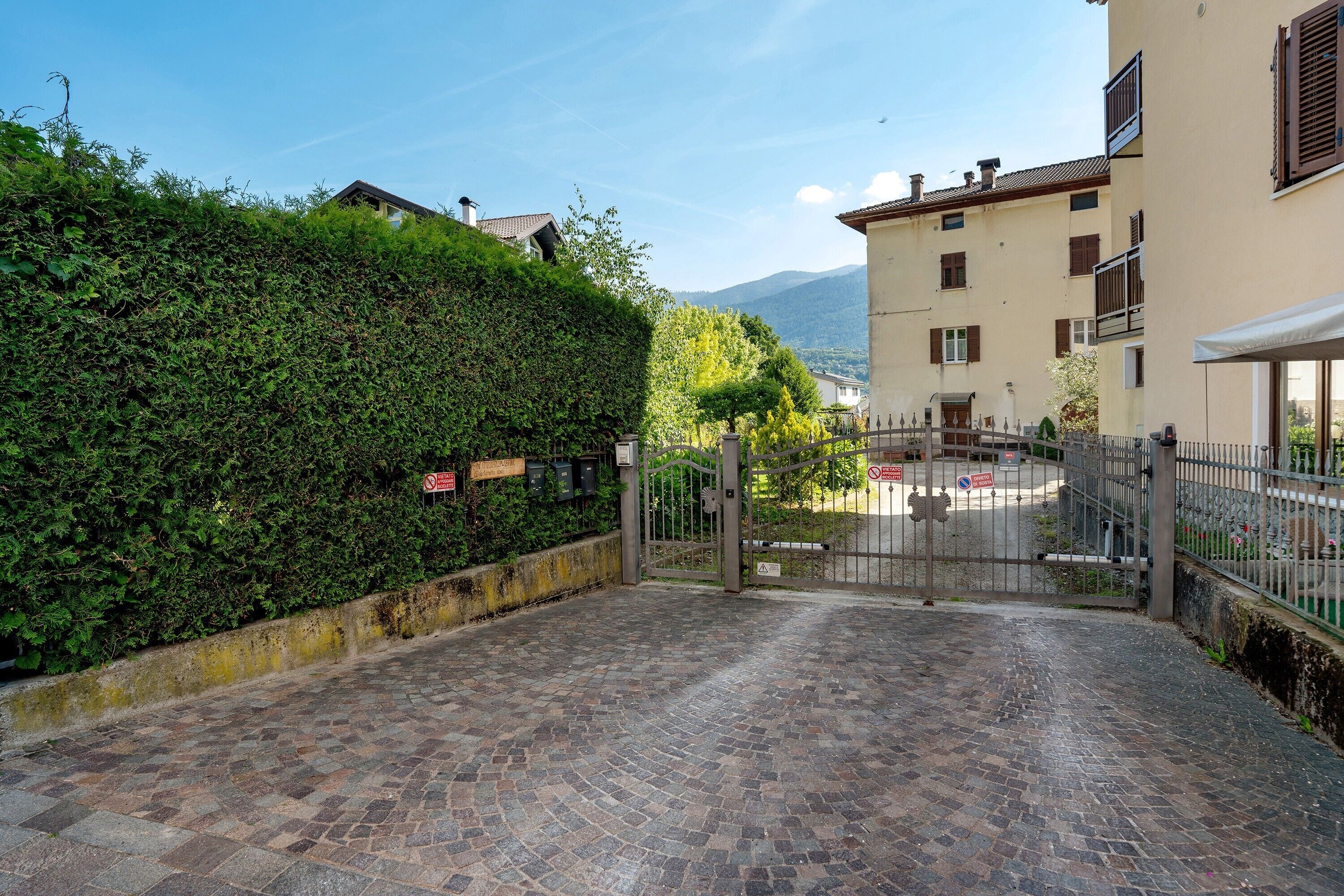 A paved entrance to a residential area with a metal gate, a hedge, and buildings with balconies.