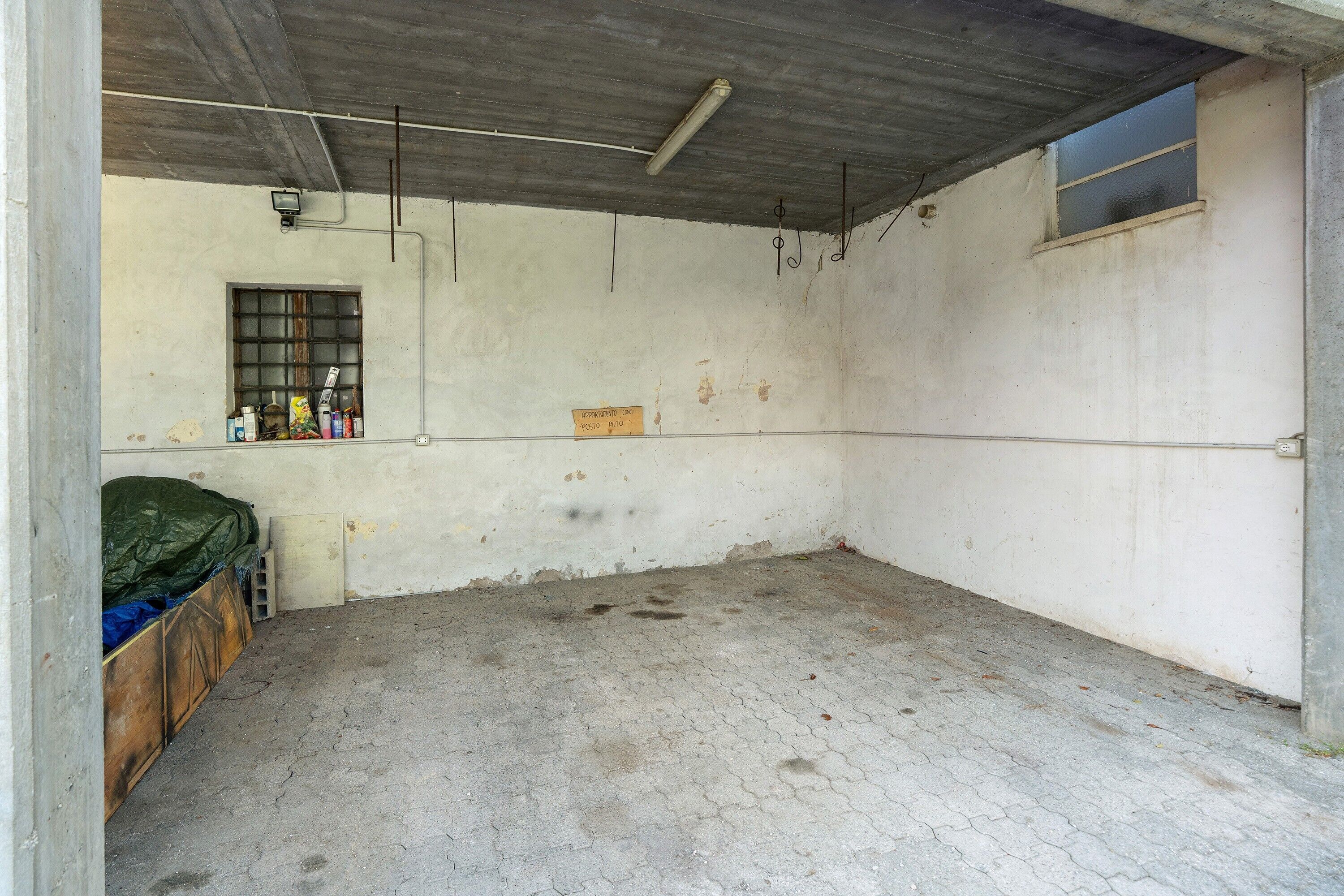 A room with a concrete floor, a window with bars, a bed with a green cover, and a small shelf with various items.