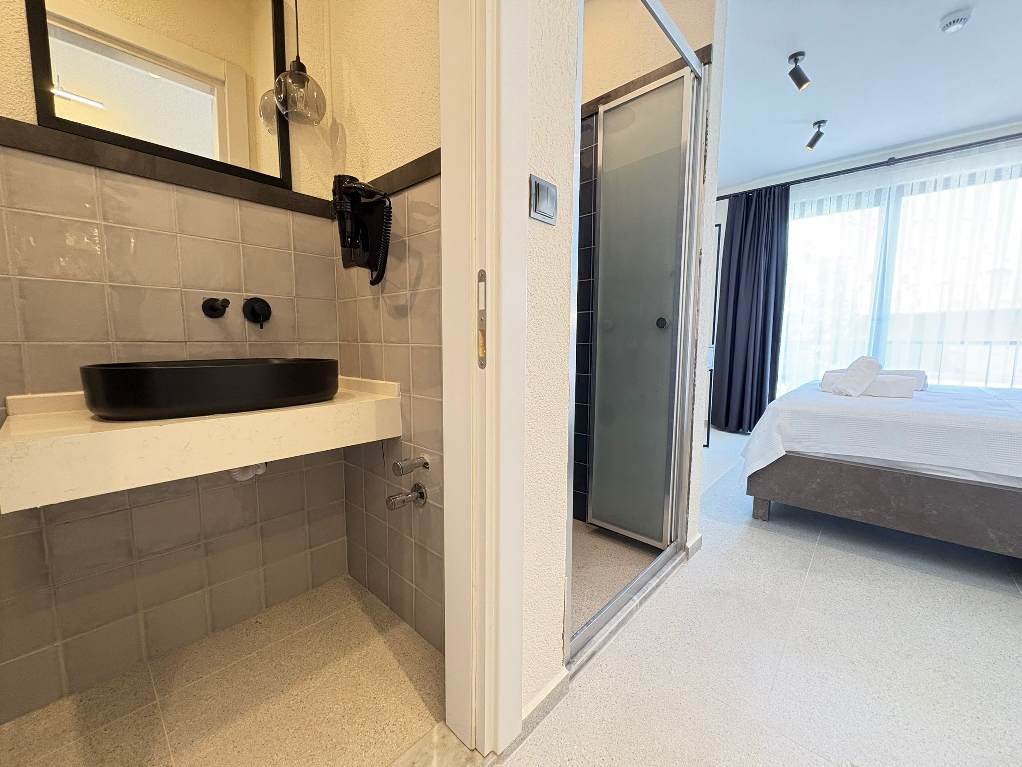 A modern bathroom with a black sink, a glass shower enclosure, and a visible door leading to a bedroom with a bed and curtains.