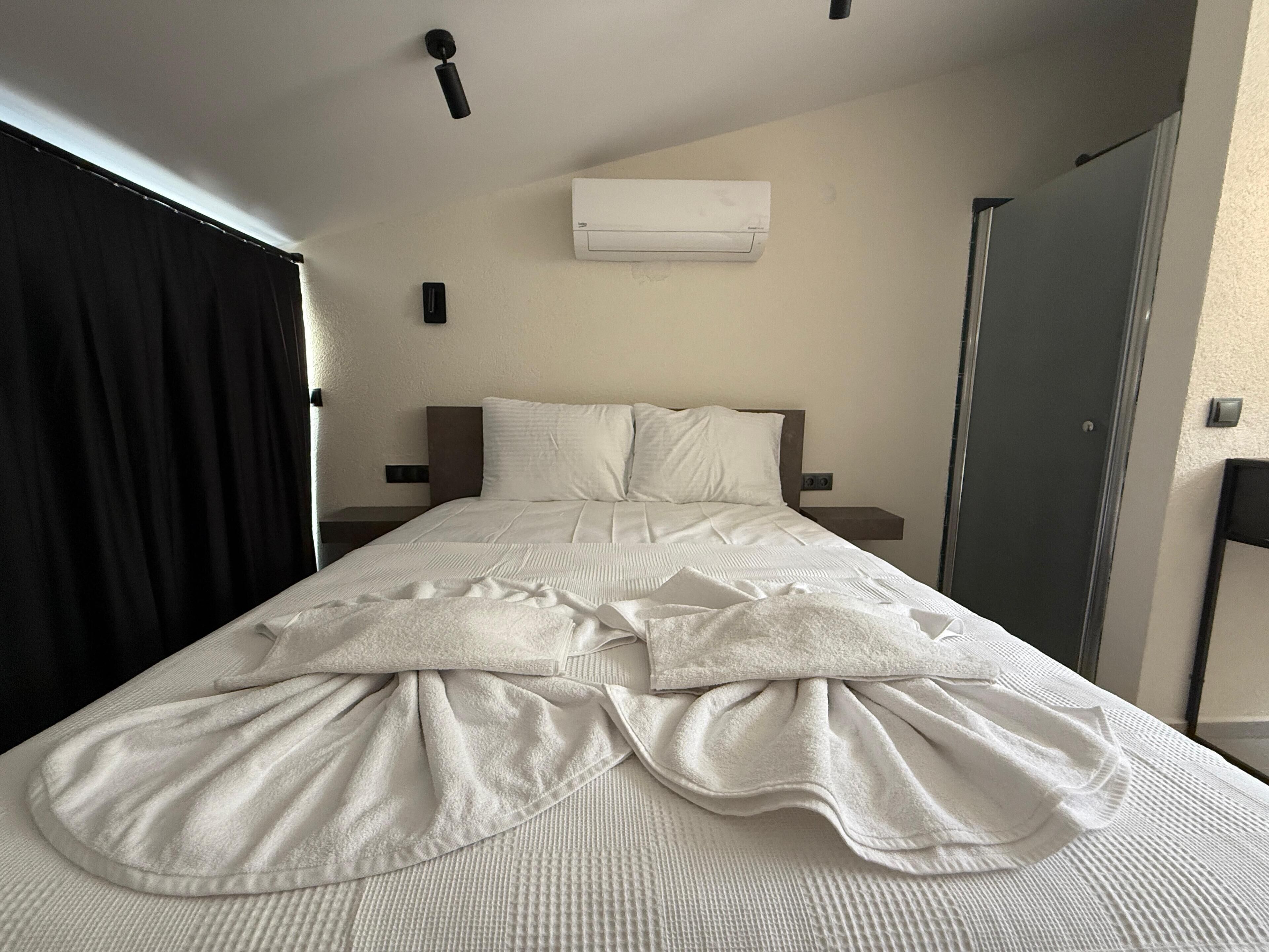 Un lit bien fait, avec une couette blanche et des oreillers, un rideau noir, un climatiseur fixé au mur et une porte sur le côté droit.